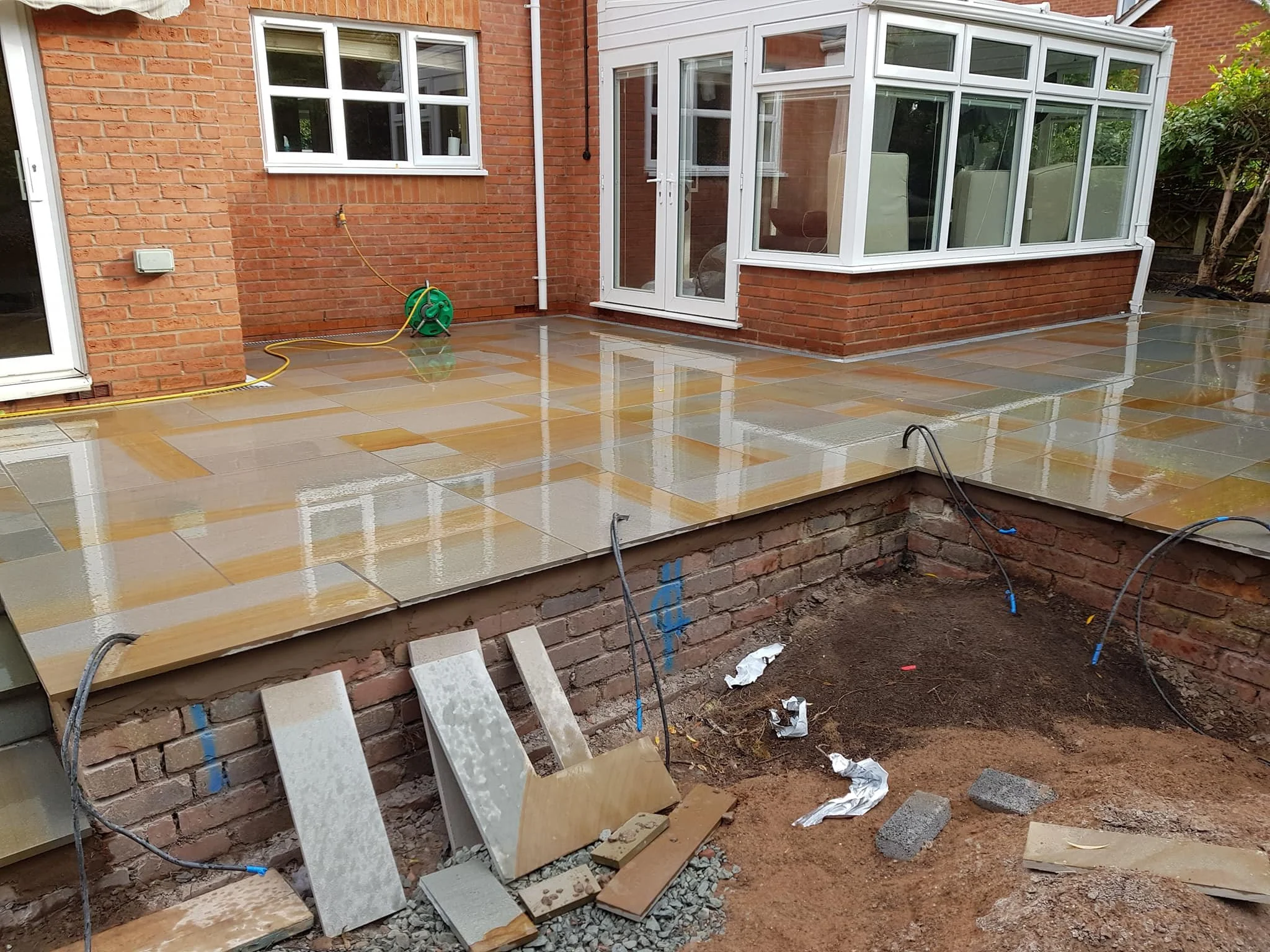 Newly constructed patio with wet tiles, adjacent to a brick house with a garden, partially bordered with brick wall, and visible construction materials and tools.