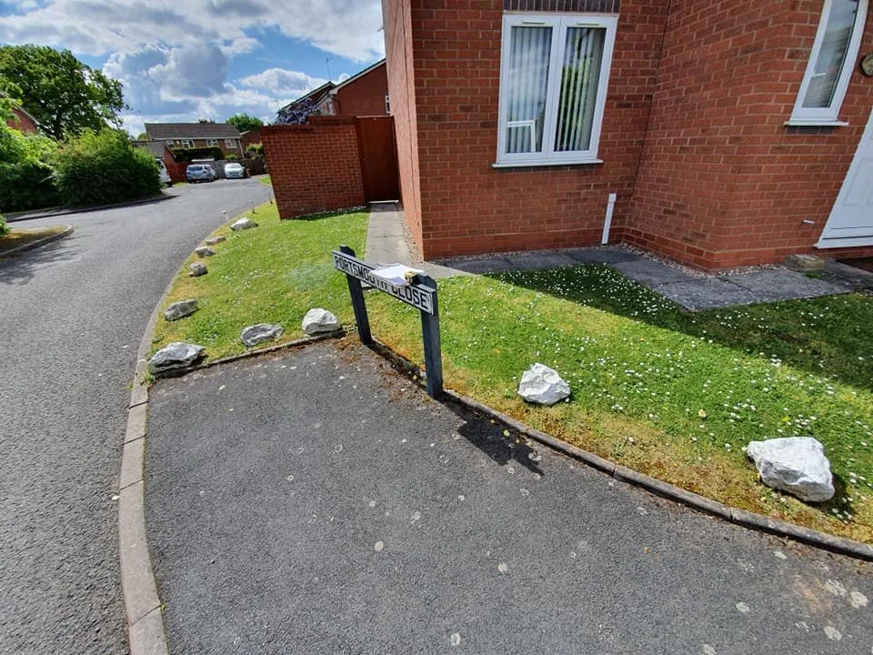 A residential street corner with a small garden area containing rocks, a sign that reads 'PRIVATE CCTV IN USE', and a red brick house with white windows.