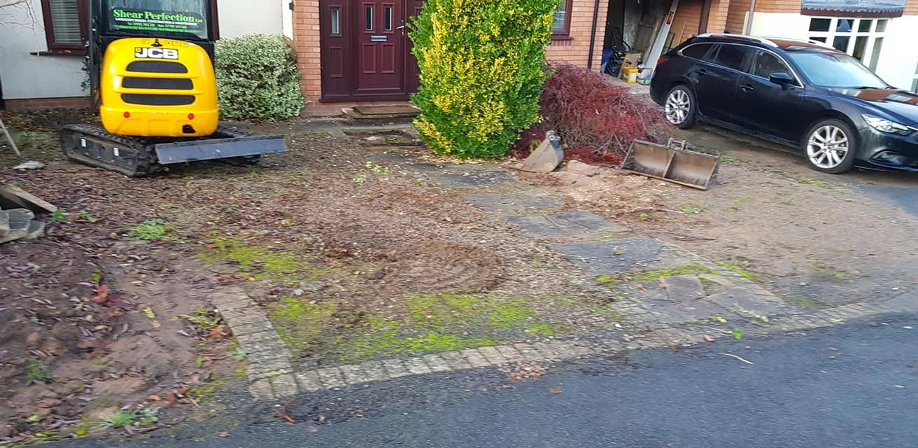 Front yard under construction with a small excavator on a dirt area, paving stones partially removed, and a car parked in the driveway.