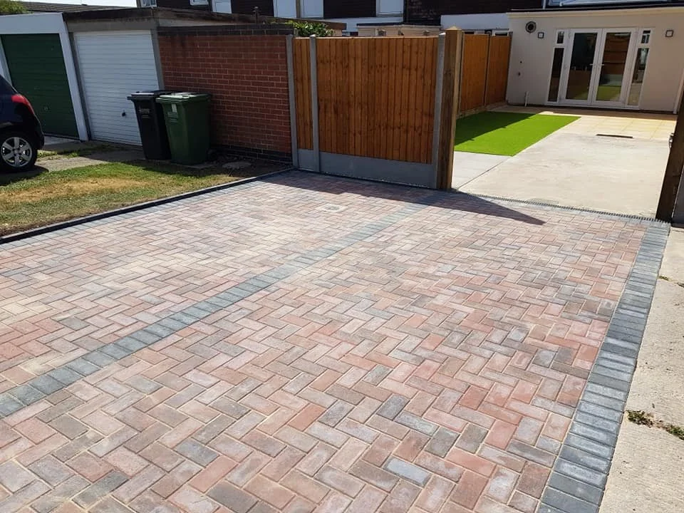 A clean brick-paved driveway leading to a gate and a backyard with a small patch of artificial grass and a patio with sliding glass doors.