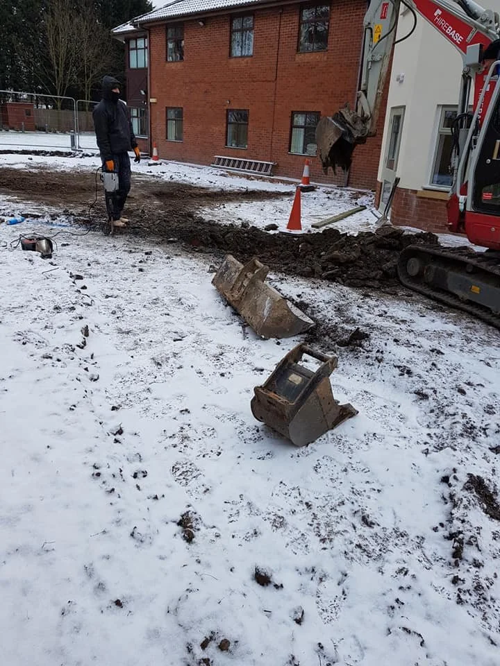 Construction site with a person operating heavy machinery, excavated trench, and fallen light fixtures in snowy ground near a brick building.