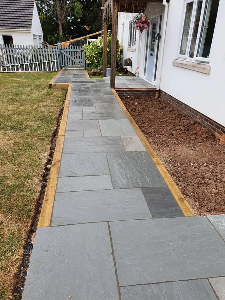 Newly paved gray stone pathway leading to a house with white exterior, a flower hanging basket, and a garden gate in the background.