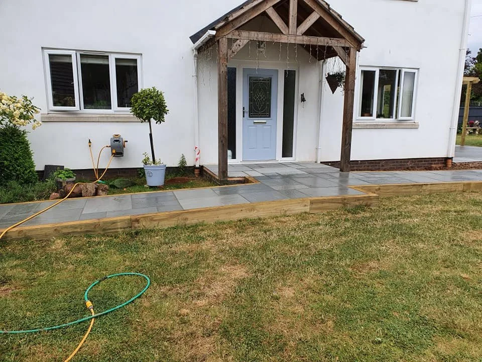 Front yard with a newly built stone walkway leading to a house porch, a small tree in a pot, a garden hose on the grass, and a white exterior wall with windows.