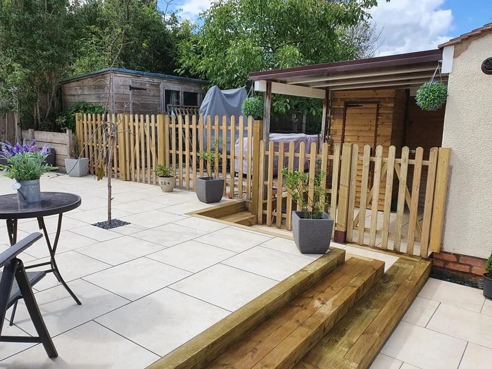 A backyard patio with light-colored tiled flooring, potted plants, a small tree, wooden stairs, and a wooden fence. There is a covered section with a hammock and decorative hanging plants. A garden shed is visible in the background, surrounded by tre