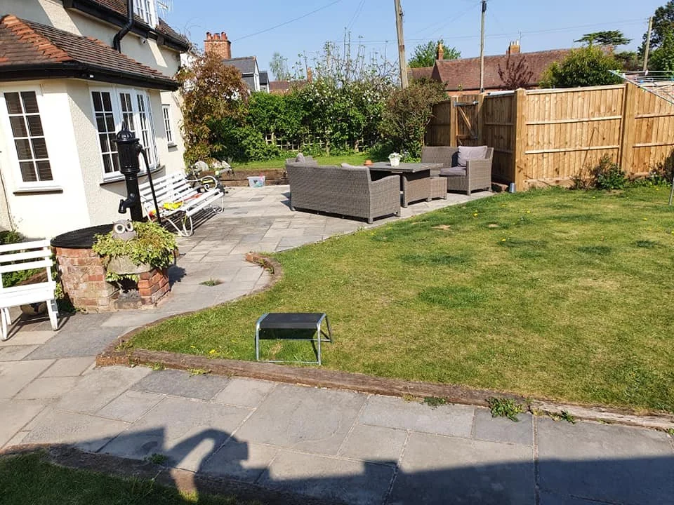 A backyard patio with a patio set of wicker couches and a table, surrounded by a stone pathway, grass lawn, a white house, benches, a garden, and a wooden fence in the background.