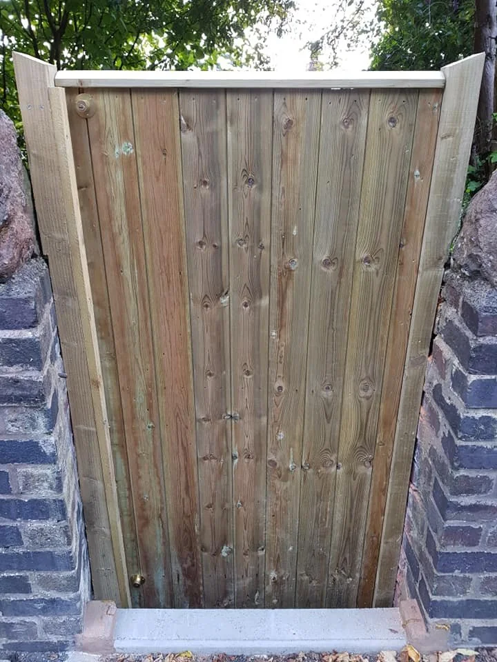 A wooden gate between brick pillars, with greenery and trees in the background.