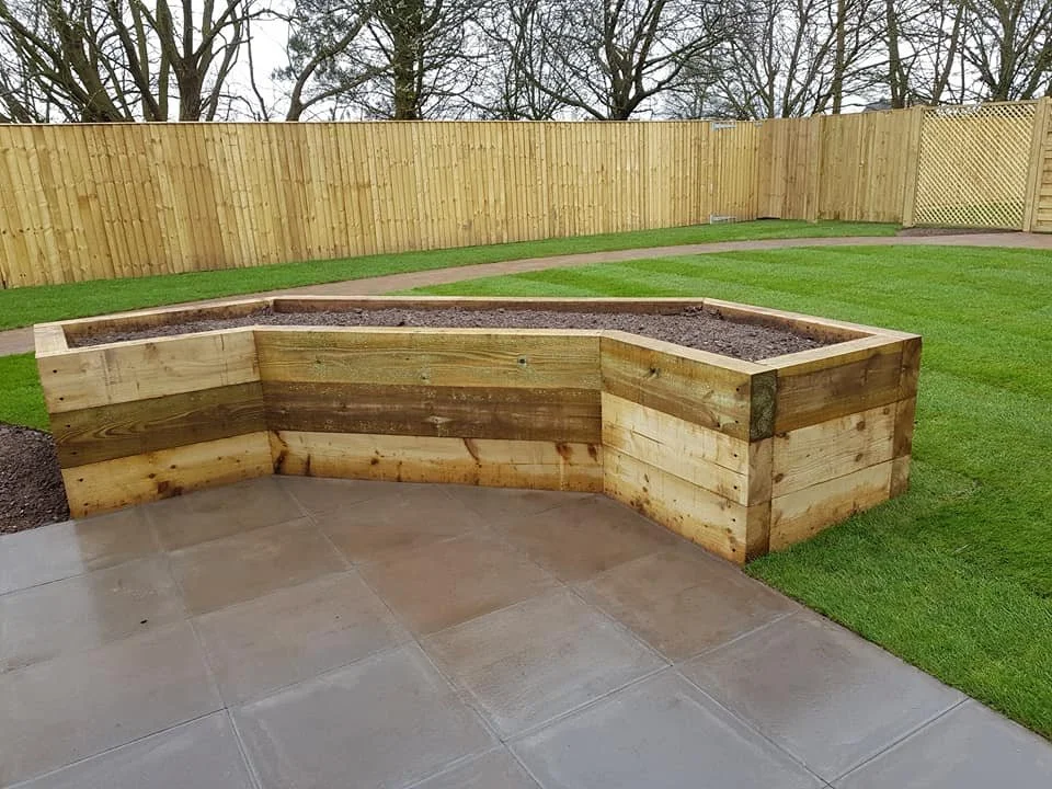 A newly built wooden raised garden bed in a backyard, with a stone patio, green grass, and a wooden fence in the background.