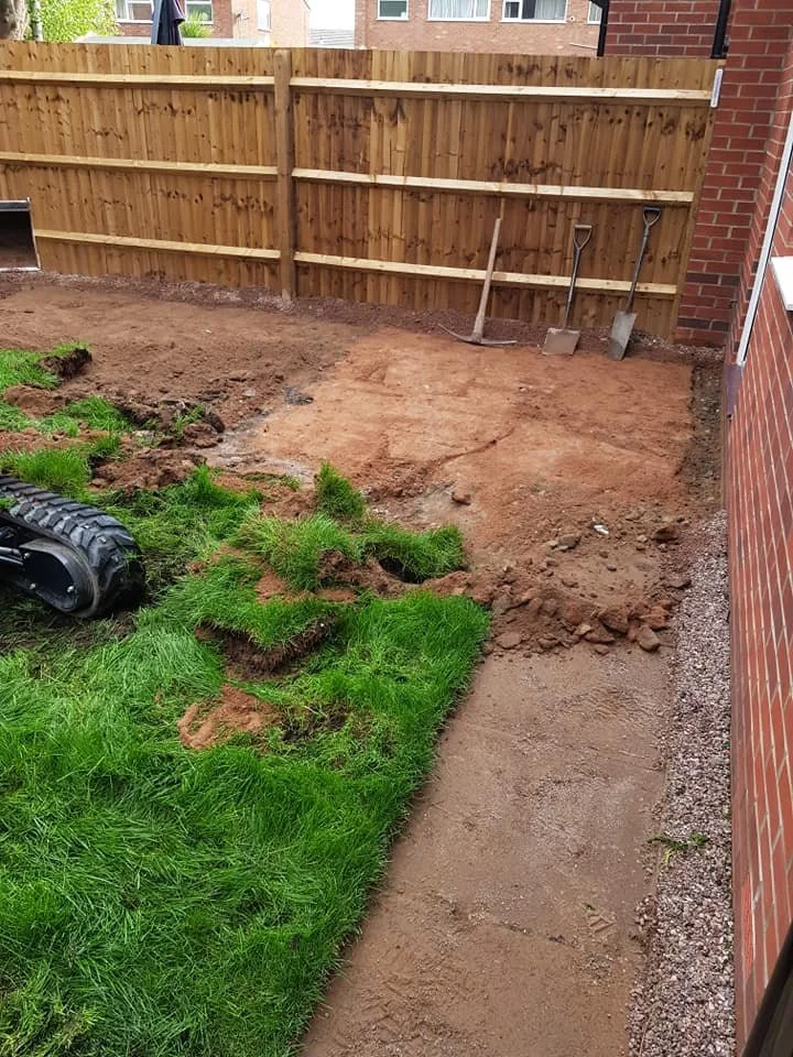 A small backyard with a dirt area being prepared for landscaping, a wooden privacy fence, a brick house wall, some gardening tools, and a patch of green grass.