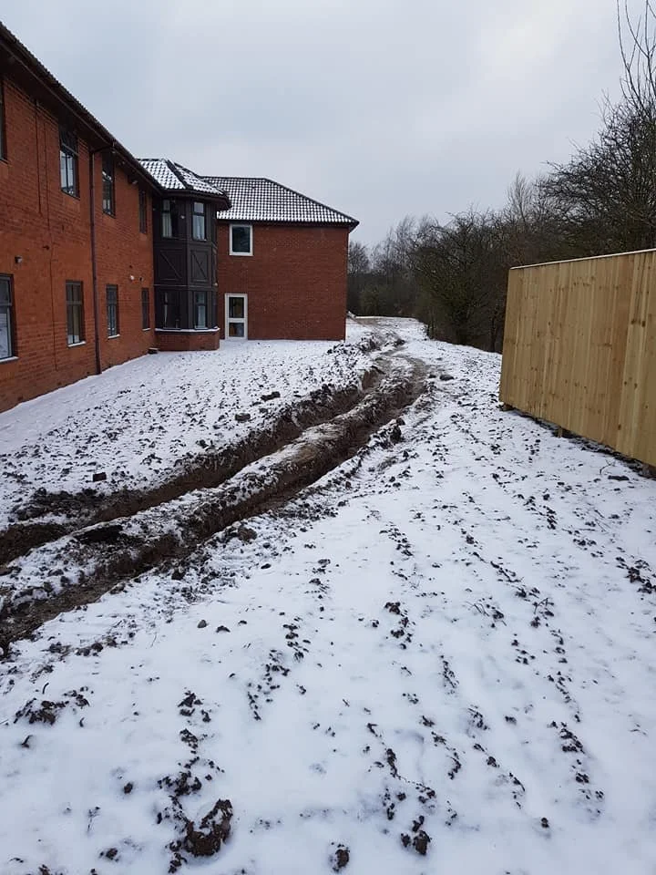 Snow-covered backyard with tire tracks leading away from a brick house with black framing, next to a wooden fence on a cloudy day.