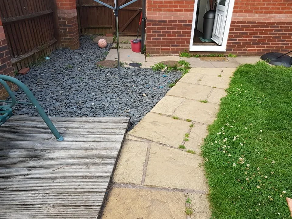 Backyard with a wooden deck, gravel area, concrete stepping stones, and a grassy patch with small white flowers. There is patio furniture, a basketball, a red bucket, and a clothesline pole.