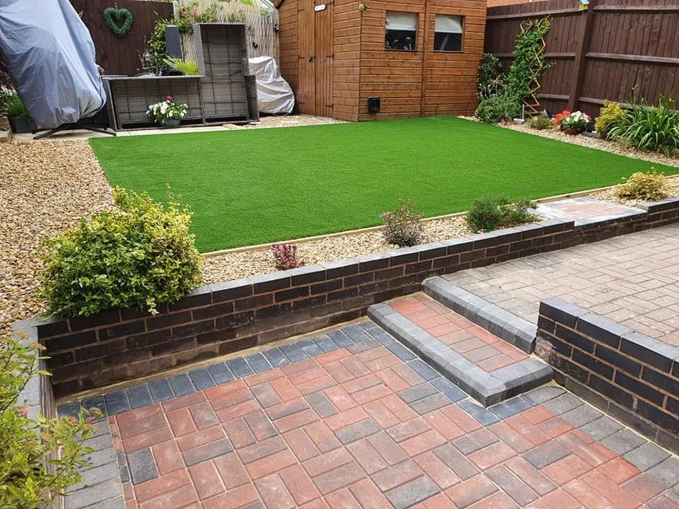A backyard garden with a small artificial grass lawn, bordered by gravel and plants, with a brick-paved patio in the foreground. There's a wooden shed and a fenced garden area in the background.