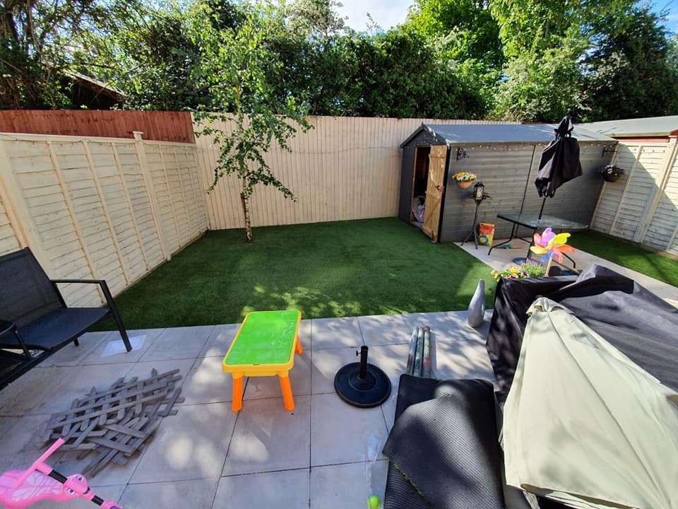 Backyard with artificial grass, a small shed, patio furniture, a children's plastic table, a folding chair, a pink tricycle, and a fenced yard with trees.