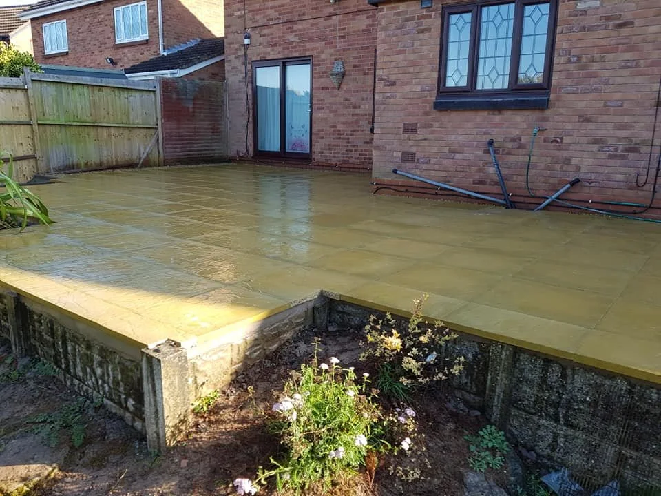 A newly installed yellow wooden deck in a backyard, with some plants and a brick house with a sliding door and window in the background.