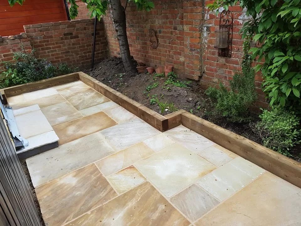Partially completed outdoor patio with sandstone tiles, wooden border, garden bed, brick wall, plants, and some garden decor in the background.