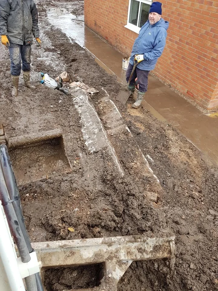 Two men working on laying concrete outside a brick building, with one shoveling wet concrete and the other standing nearby.