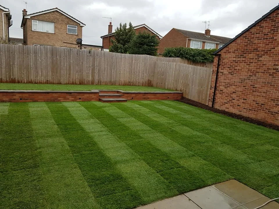A backyard with a freshly mowed lawn, striped with alternating light and dark green grass. There are wooden steps leading up to an elevated area with a wooden deck, bordered by a wooden fence. The sky is overcast, and neighboring houses are visible i