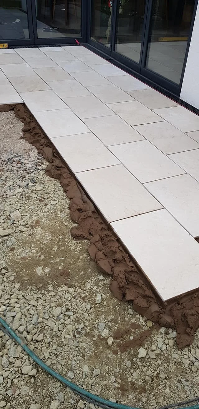 Newly tiled outdoor patio area with large gray tiles next to a glass sliding door, with soil and gravel underneath.