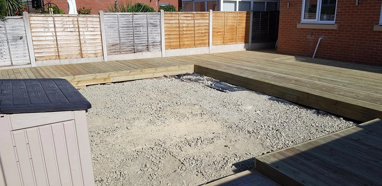 A backyard under construction with a gravel base, wooden decks, and a multi-colored wooden privacy fence. A house with brick exterior is visible on the right.