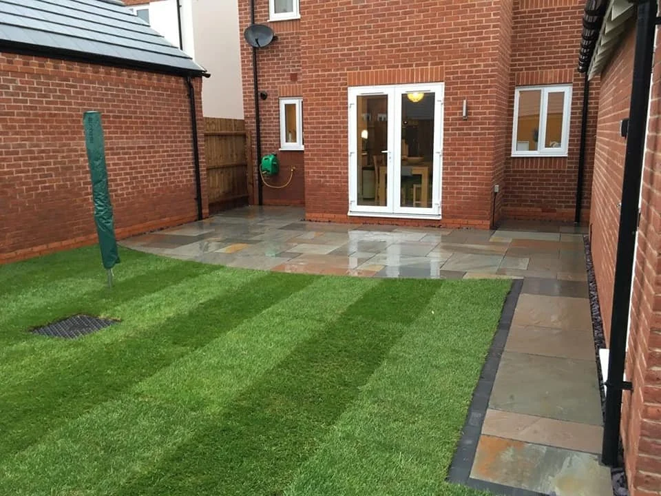 A backyard with freshly laid green grass patches, a wet stone patio, and a sliding glass door leading into the house. The yard is enclosed with brick walls and has a covered clothesline.