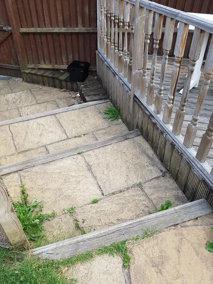 A small outdoor patio area with worn stone paving and wooden railings, some weeds growing between the stones, and a black storage container in the corner.