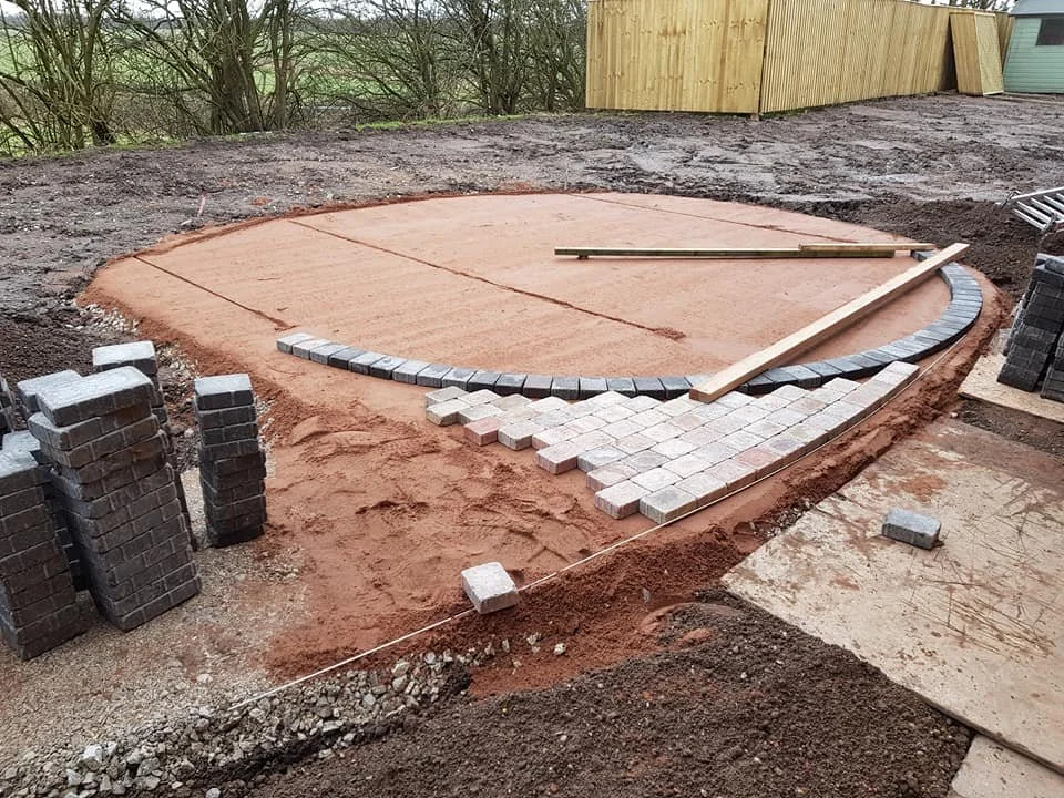 Construction site with a new circular patio area being built using grey and red bricks, surrounded by dirt and construction materials, with a wooden fence in the background.