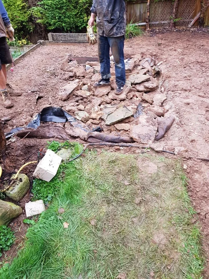 A person working on a garden or yard project, standing on a pile of rocks and dirt, with some tools and equipment nearby. Part of a grassy area is in the foreground, and a fence with greenery behind.