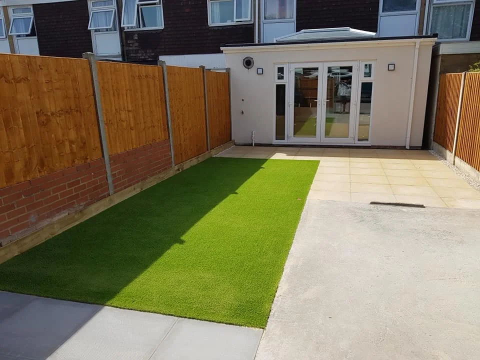 Backyard with a small green artificial lawn, concrete patio, wooden fence, and glass double doors leading to an interior room.