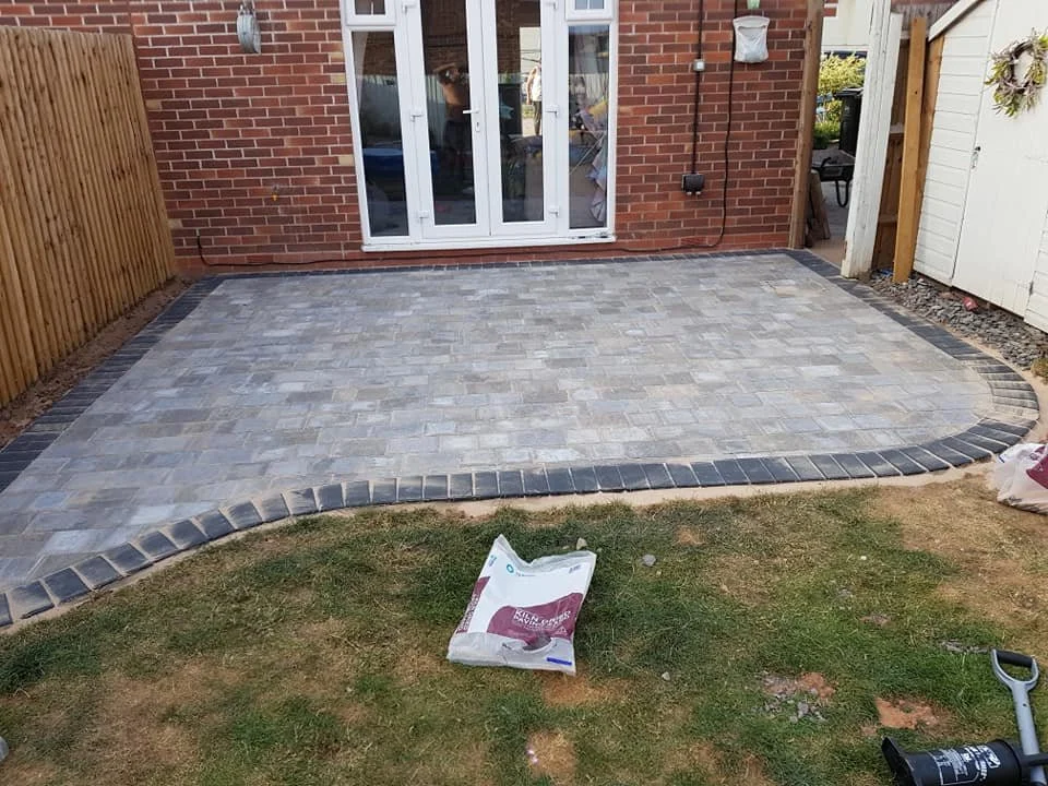 A newly constructed brick patio with a brick border in a backyard, adjacent to a house with white sliding doors and outdoor fencing.