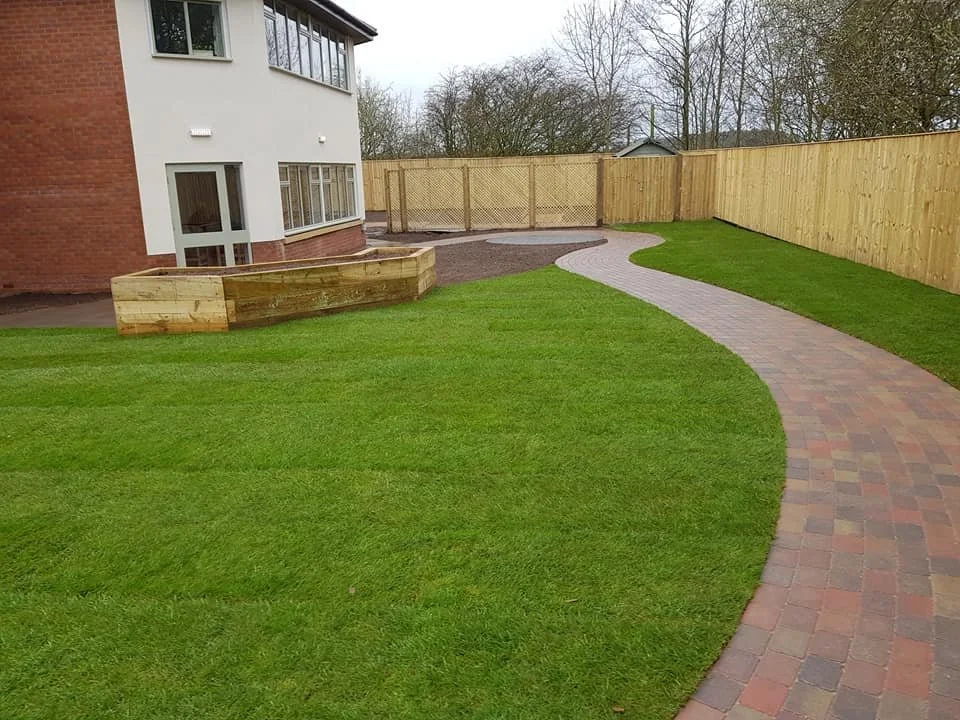 A backyard with a curved brick pathway, a grassy lawn, a wooden fence, and a multi-story house with a brick and white exterior.