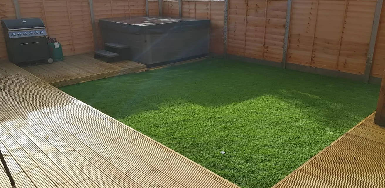 A backyard with a wooden deck, green artificial grass, a hot tub, and a barbecue grill.