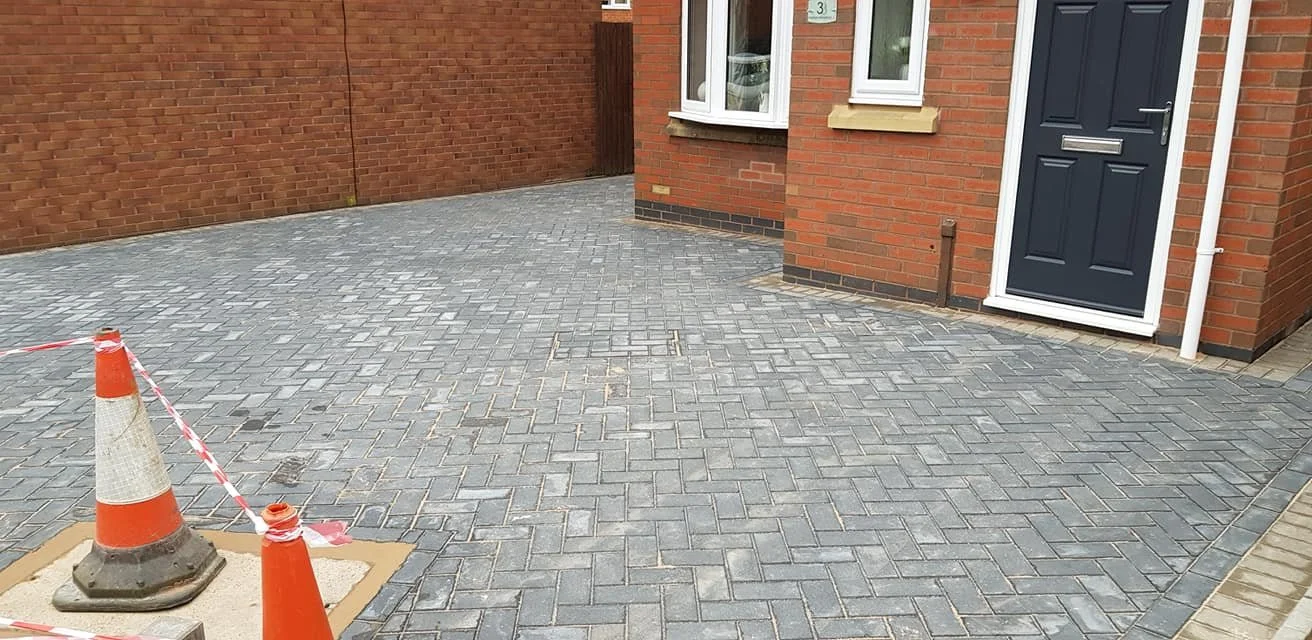 Freshly paved driveway with orange cones and caution tape, located outside a brick house with a blue front door and white window frames.