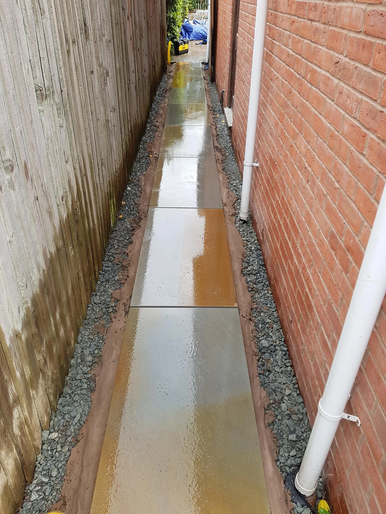 Wet concrete pathway between a wooden fence on the left and a brick wall on the right, with a white drainpipe attached to the wall, leading to a backyard with some equipment and tarps.