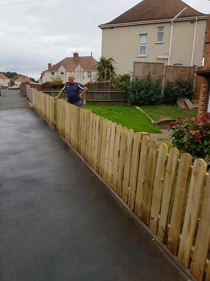 A man standing behind a newly installed wooden fence along a residential street, with houses and a partly cloudy sky in the background.