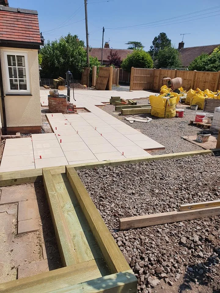 Construction site with paving stones being laid for a sidewalk or pathway next to a house, with building materials and tools visible, and a wooden fence in the background.