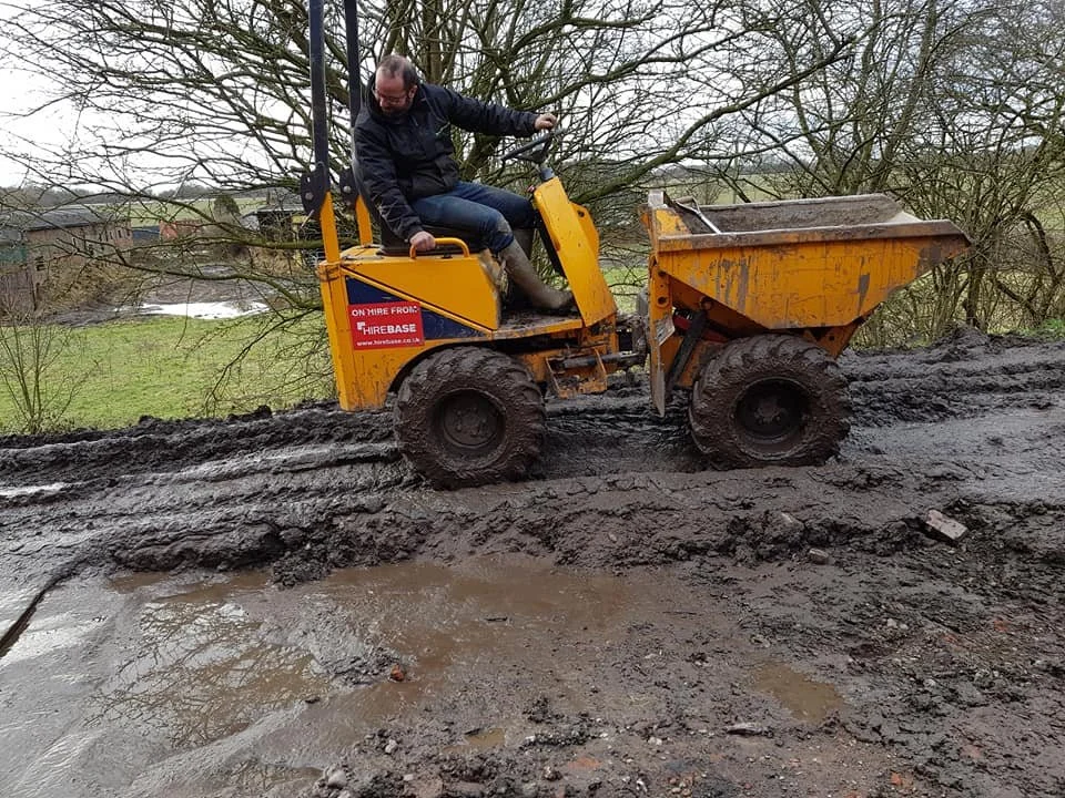 Man operating a small yellow construction vehicle with a rear dump bucket on muddy terrain near trees and a grassy area.