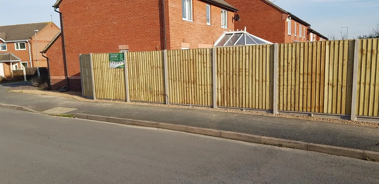 Residential street with newly installed wooden fence along sidewalk, brick house in background, and a small gate in the fence.