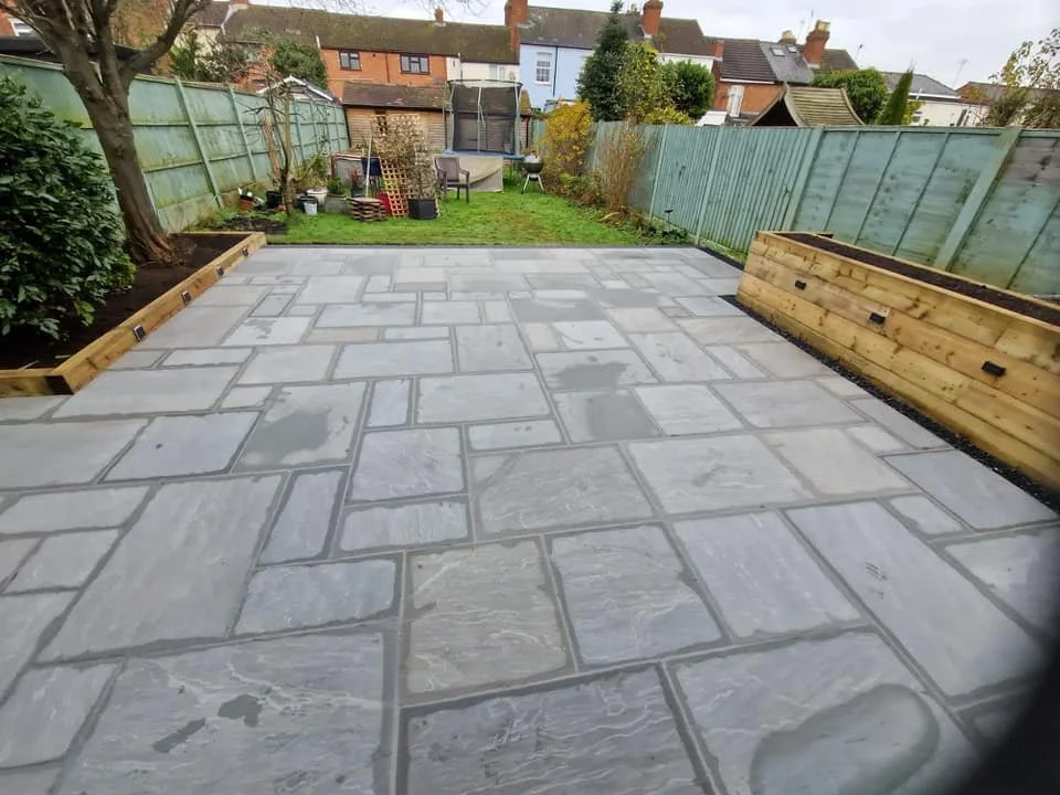 A newly paved backyard patio with large gray stone tiles, wooden planter boxes along the sides, and a fenced yard with a green lawn, trees, and garden furniture in the background.
