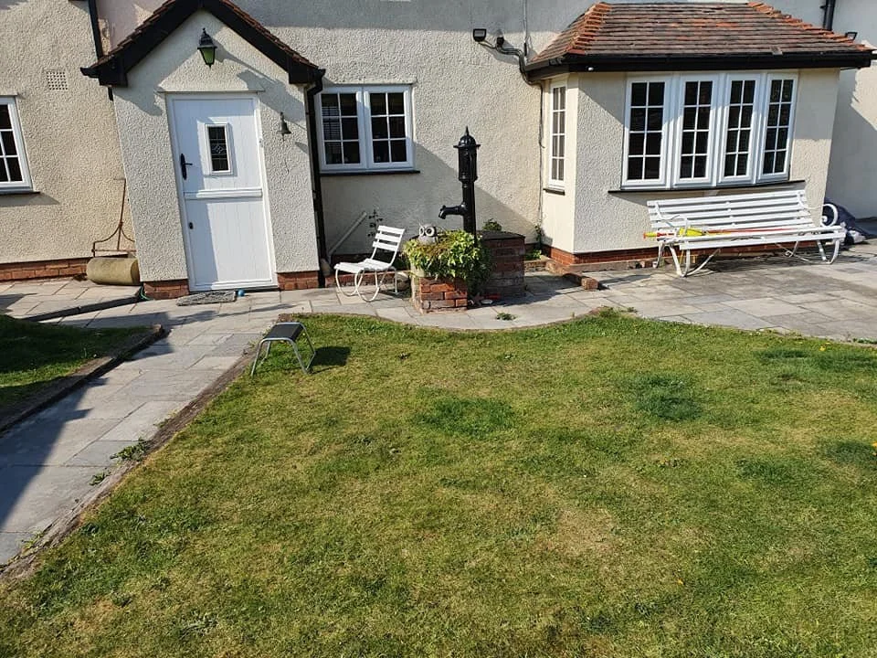 View of the back yard of a house with a small lawn, a paved patio, white outdoor furniture, a water pump, and a white door on the house exterior.