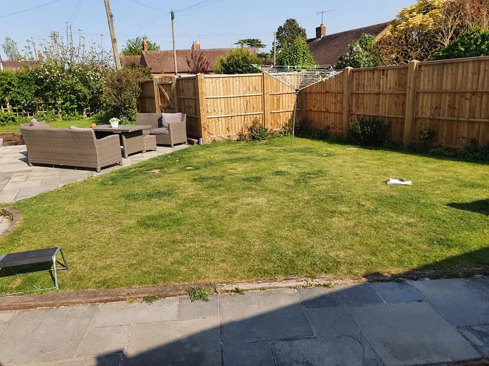 A backyard with a grassy lawn, a wooden fence, a patio area with outdoor furniture, a rotary clothesline, and trees and houses in the background under a clear blue sky.