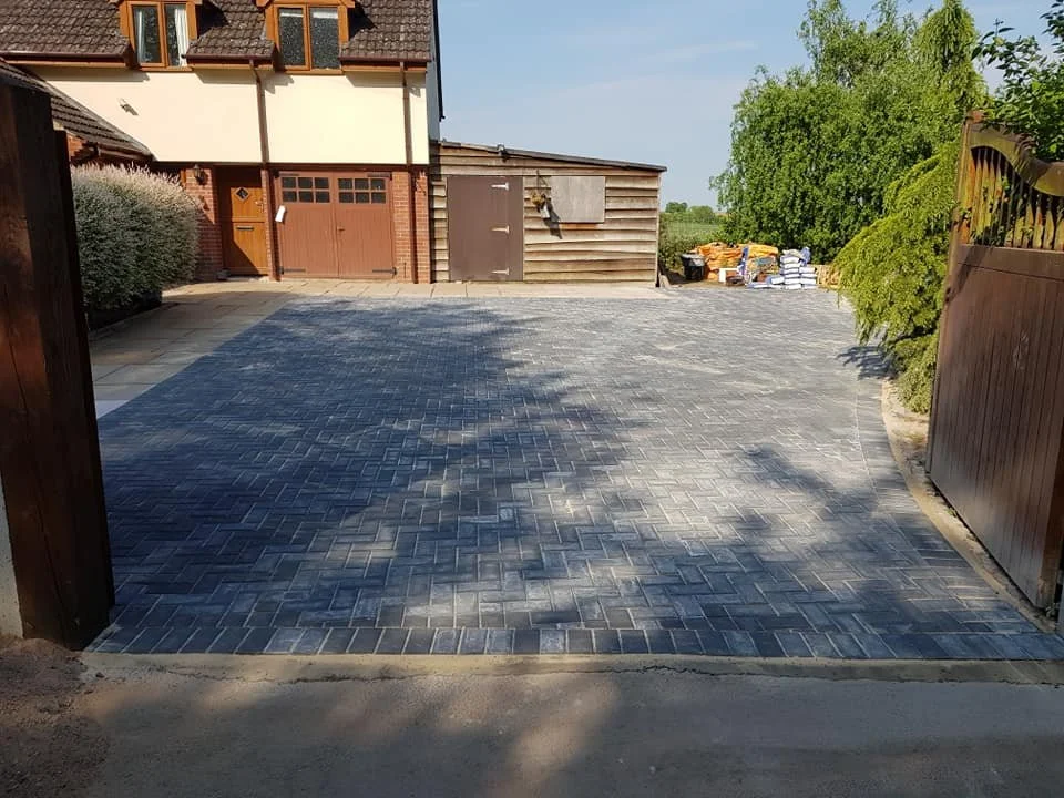 A newly paved driveway with dark-colored bricks leading to a house with a garage and wooden exterior. There are trees and shrubs on the sides, with some construction materials visible in the background.
