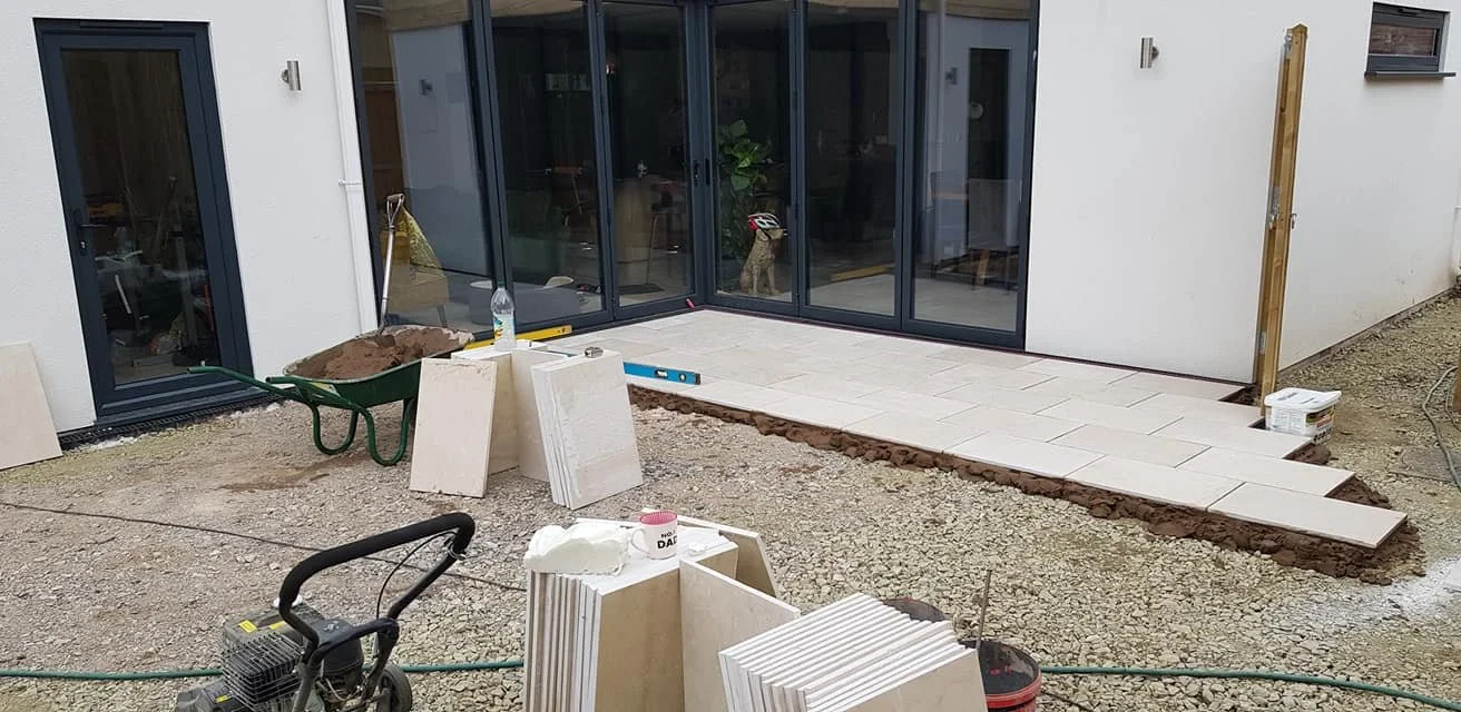 Construction site for a tiled patio outside a modern house with large glass sliding doors. Paving stones are partially laid, and tools and materials are scattered around.
