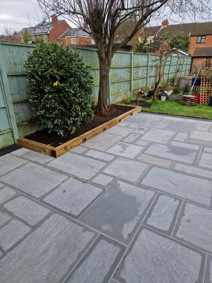 Backyard with stone patio, a raised flower bed with dark soil, a large tree, a green shrub, and a wooden fence in the background.