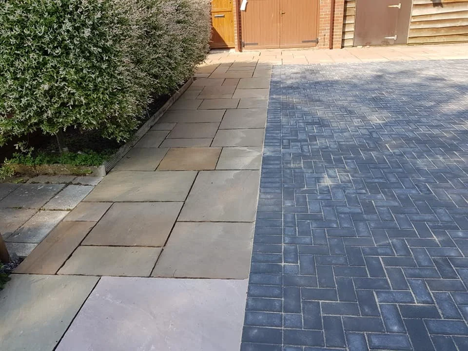 Sidewalk with two different types of paving: beige stone slabs on the left and dark gray bricks on the right, near a wooden fence and some bushes.