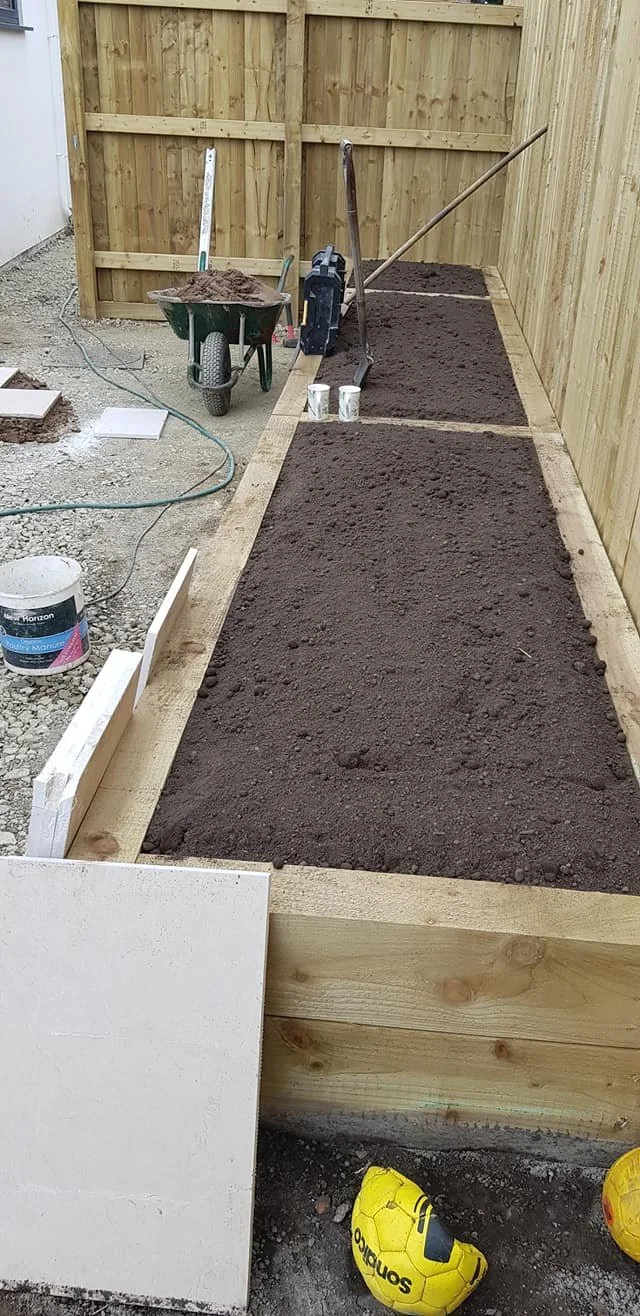 A small garden bed under construction with fresh soil, surrounded by a wooden frame, with gardening tools and supplies nearby.