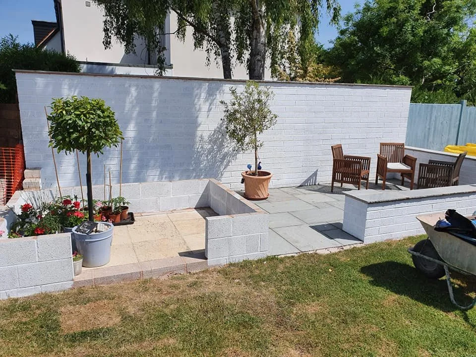 A backyard patio with a white brick wall, potted plants, and outdoor furniture including a table and four chairs, with a wheelbarrow on the grass in the foreground.