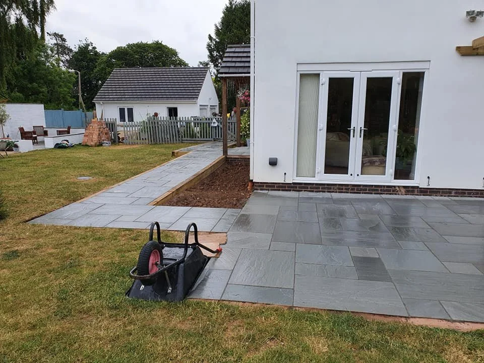 Backyard with newly laid gray stone patio, a wheelbarrow, and a white house with a glass door and brick base, overcast sky.