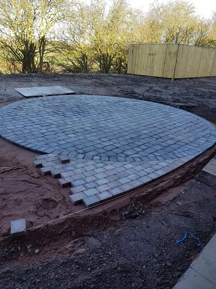 Undergoing paving, a curved area with gray stone pavers, with some bricks and sand around it, and a wooden fence and trees in the background.