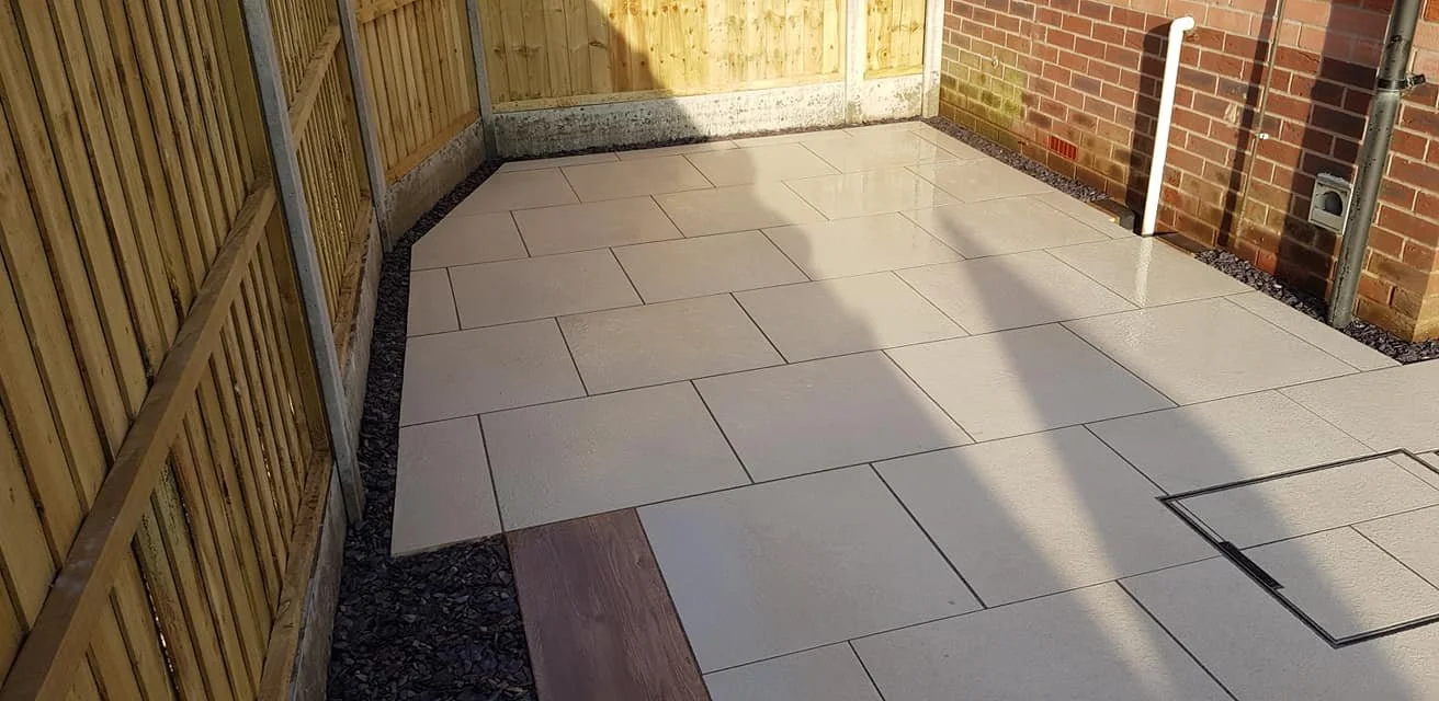 A newly paved backyard patio with large rectangular beige tiles, a wooden fence on the left, and a brick wall on the right. There's a white pipe attached to the brick wall and a drainage grate on the tiled surface.