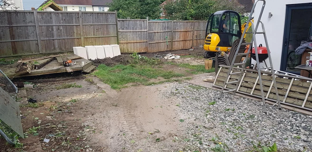 Backyard undergoing landscaping or construction with gravel, dirt, and construction tools, including a small yellow excavator, a ladder, stacked wooden planks, and potted plants.