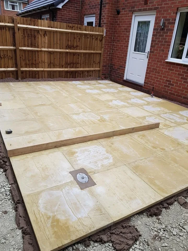 Backyard patio with newly laid beige stone tiles, partially frosted with floor leveling compound, enclosed by a wooden fence and a brick house with a white door and window.